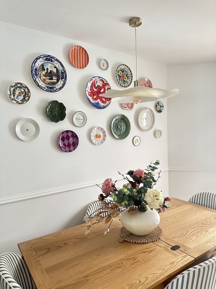 Parede com composição de pratos decorativos sobre mesa de madeira com arranjo de flores e pendente de tecido
